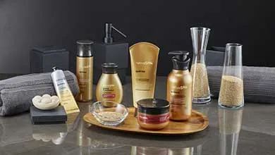 Gold and black spa products, rolled grey towels, a wooden tray, and decorative glass containers arranged on a dark, reflective surface.