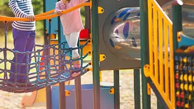 Two young children cross a blue rope bridge on a colorful playground.