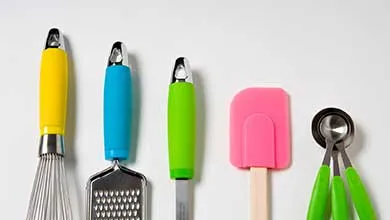 Colorful kitchen utensils—a whisk, grater, peeler, spatula, and measuring spoons—arranged in a row on a white background.