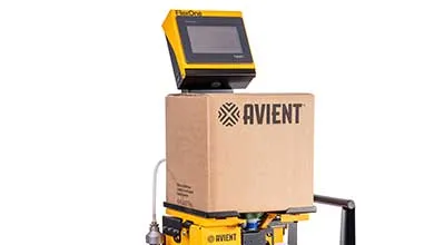 A yellow industrial machine holds a cardboard box labeled "AVIENT," with a control panel and screen placed on top of the box.