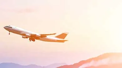 A commercial airplane lifts off from a wide airport runway at sunrise, with runway lights glowing in the foreground and layered blue mountains silhouetted against a pink‑and‑gold sky in the background.