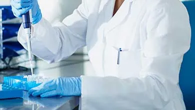 Laboratory professional using a pipette to transfer liquid into sample wells on a lab bench with bottles and equipment nearby.