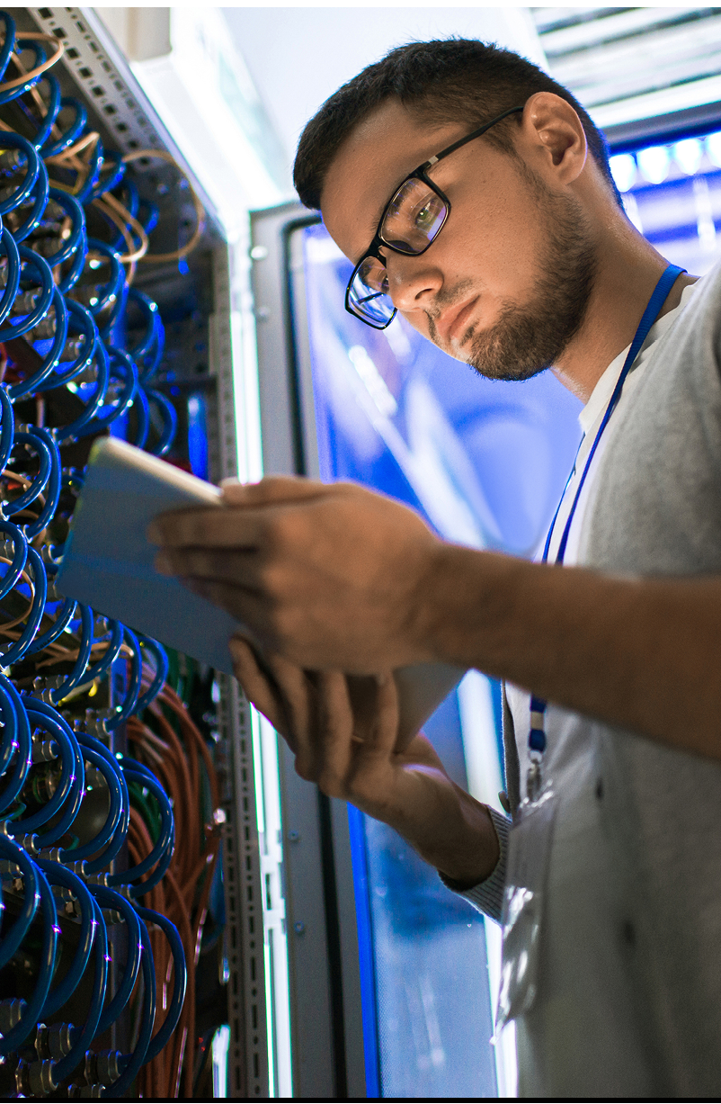 Low angle portrait of young man using digital tablet standing by server cabinet while working with supercomputer in blue light